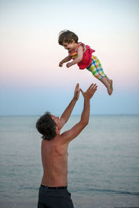 Shirtless father carrying daughter while standing at beach against sky