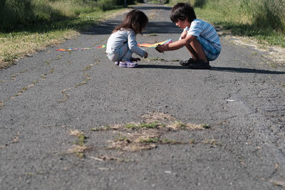 Siblings with kite crouching on road during sunny day