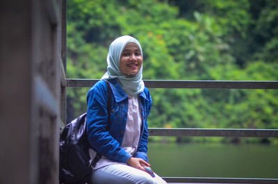 Portrait of smiling young woman sitting by railing