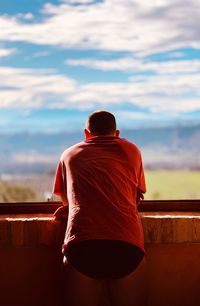 Rear view of man leaning on retaining wall against cloudy sky