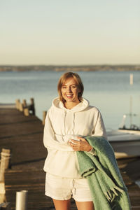 Portrait of smiling young woman holding blanket while standing at jetty