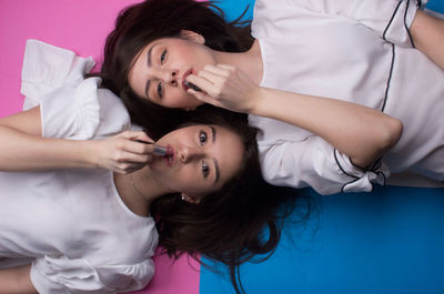Directly above shot of female friends applying lipstick while lying on colored background