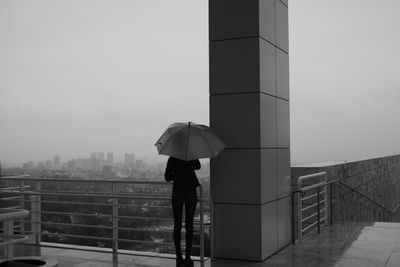 Rear view of woman with umbrella standing by railing on building during rainy season