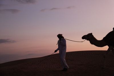 Man with camel at desert against sky during sunset