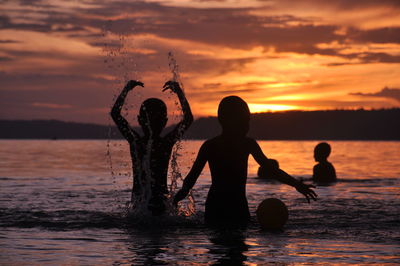 Silhouette children playing in sea against sky during sunset
