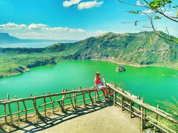 Woman looking at lake while sitting on railing during sunny day