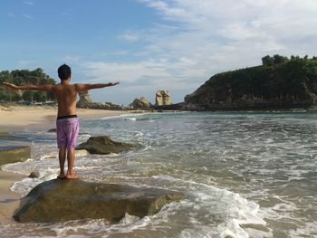 Rear view of shirtless man with arms outstretched standing on rock at sea 