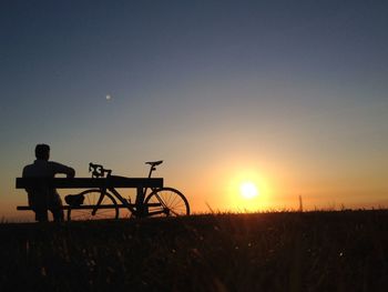 Rear view of man sitting on bench by bicycle at field against sky during sunset