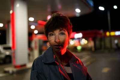 Lonely millennial woman in gray coat poses stands near with night gas station waiting for friends