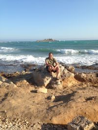 Portrait of mature man sitting on rock at beach against clear sky