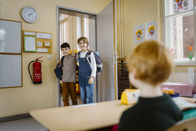 Happy schoolboys with arms around entering classroom and looking at friend