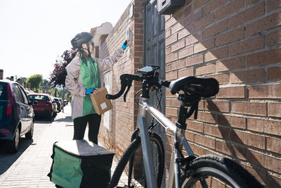 Female courier with parcel pressing button on intercom device on brick wall while making delivery on sunny day in town
