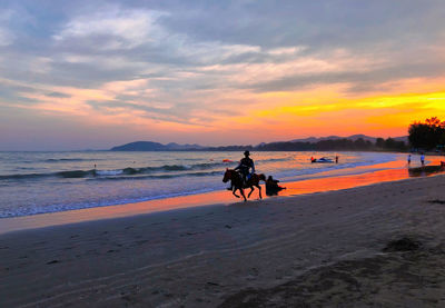 People riding horse at beach against sky during sunset
