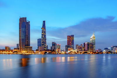 Illuminated urban skyline by river against sky during sunset
