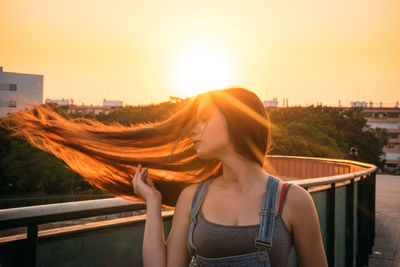 Young woman standing by railing against sunset in city