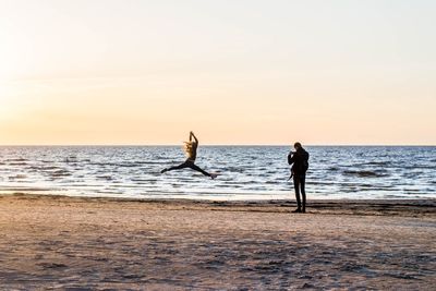 Man photographing female friend jumping at beach during sunset