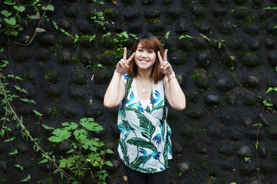 Portrait of cheerful young woman gesturing peace sign while standing by wall