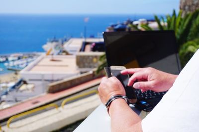 Cropped image of person using smart phone by laptop on sunny day