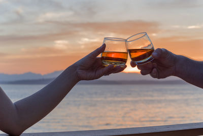 Cropped image of hands toasting against sea during sunset