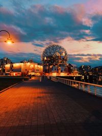 Illuminated street by buildings against sky at sunset