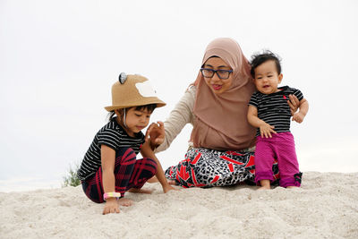 Mother with daughters sitting on sand at beach against clear sky