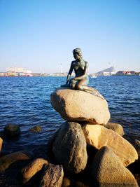 Statue of man sitting on rock by sea against clear sky