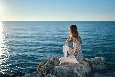 Rear view of woman sitting on rock by sea against sky