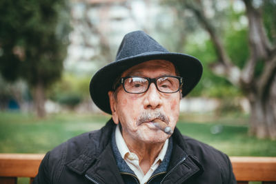 Senior male with mustache wearing hat smoking cigar and looking at camera while sitting on bench in park with green trees on blurred background