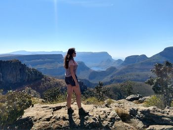 Side view young woman standing on cliff by mountains against sky