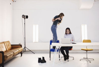 Blogger photographing female colleague using laptop at desk in creative office