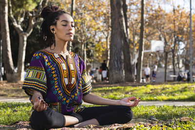 Beautiful young woman meditating while practicing lotus position yoga at park on sunny day
