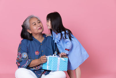 Girl kissing grandmother holding gift against pink background