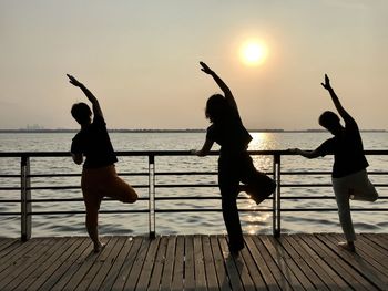 Rear view of silhouette friends exercising on pier by lake against sky during sunset