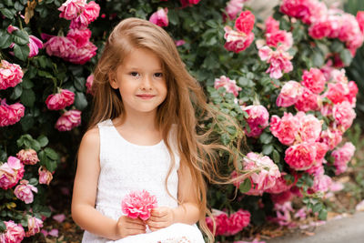 Cute baby girl 5-6 year old hold rose flower sit over bloom bushes in garden outdoor. spring season