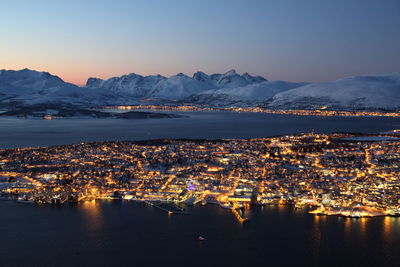 Illuminated cityscape by sea and snow covered mountains against clear sky at dusk