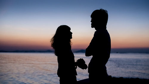 Two people standing on shore at sunset