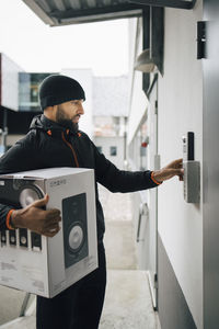 Serious male worker ringing intercom while carrying box during delivery