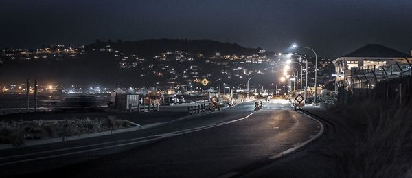 Illuminated street amidst buildings against sky at night