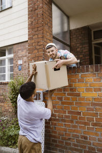 Gay couple helping each other while holding cardboard box over brick wall