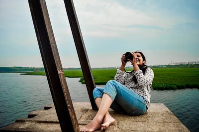 Young woman photographing through camera while sitting against lake