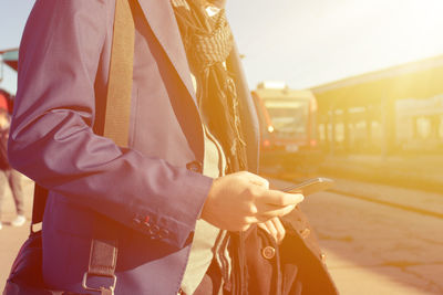 Midsection of man holding mobile phone while standing at railroad station platform