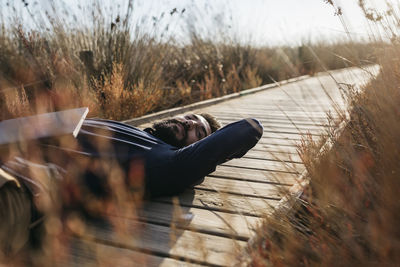 Man with laptop lying down amidst field on boardwalk during sunny day