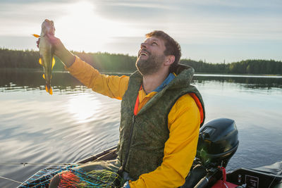 Happy man holding haddock in lake against sky