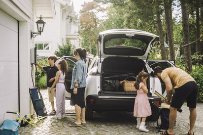 Family charging and loading luggage in trunk of electric car outside house