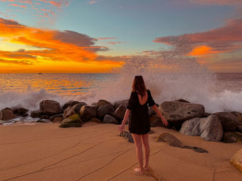Rear view of woman standing at beach against sky during sunset