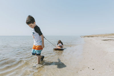 Boy pulling brother sitting on surfboard in sea against clear sky during sunny day