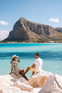 Woman sitting on rock by sea against mountains