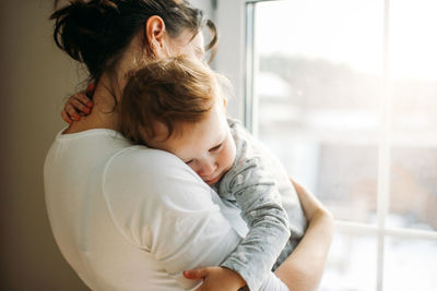 Mother embracing baby girl while standing against window