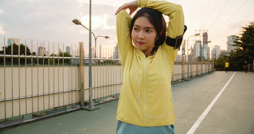 Beautiful young woman exercising while standing in city against sky