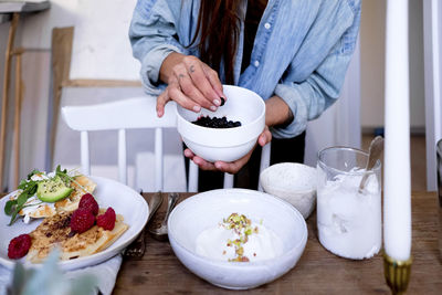 Midsection of woman adding blackberries in yogurt on plate by vase at wooden table